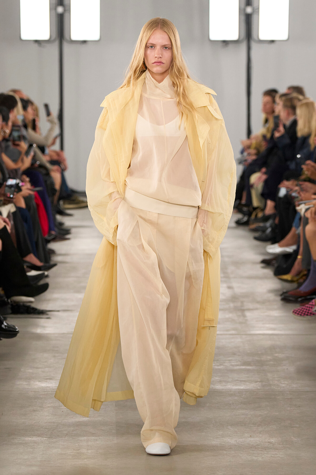 Model walking a fashion show in a sheer cream blouse and wide pale pants under a long pale-yellow coat, runway audience on both sides.