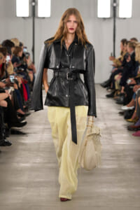 Model walks the runway in a cropped black leather belted jacket with a cape over pale yellow wide-leg pants, carrying a cream fringe bag.