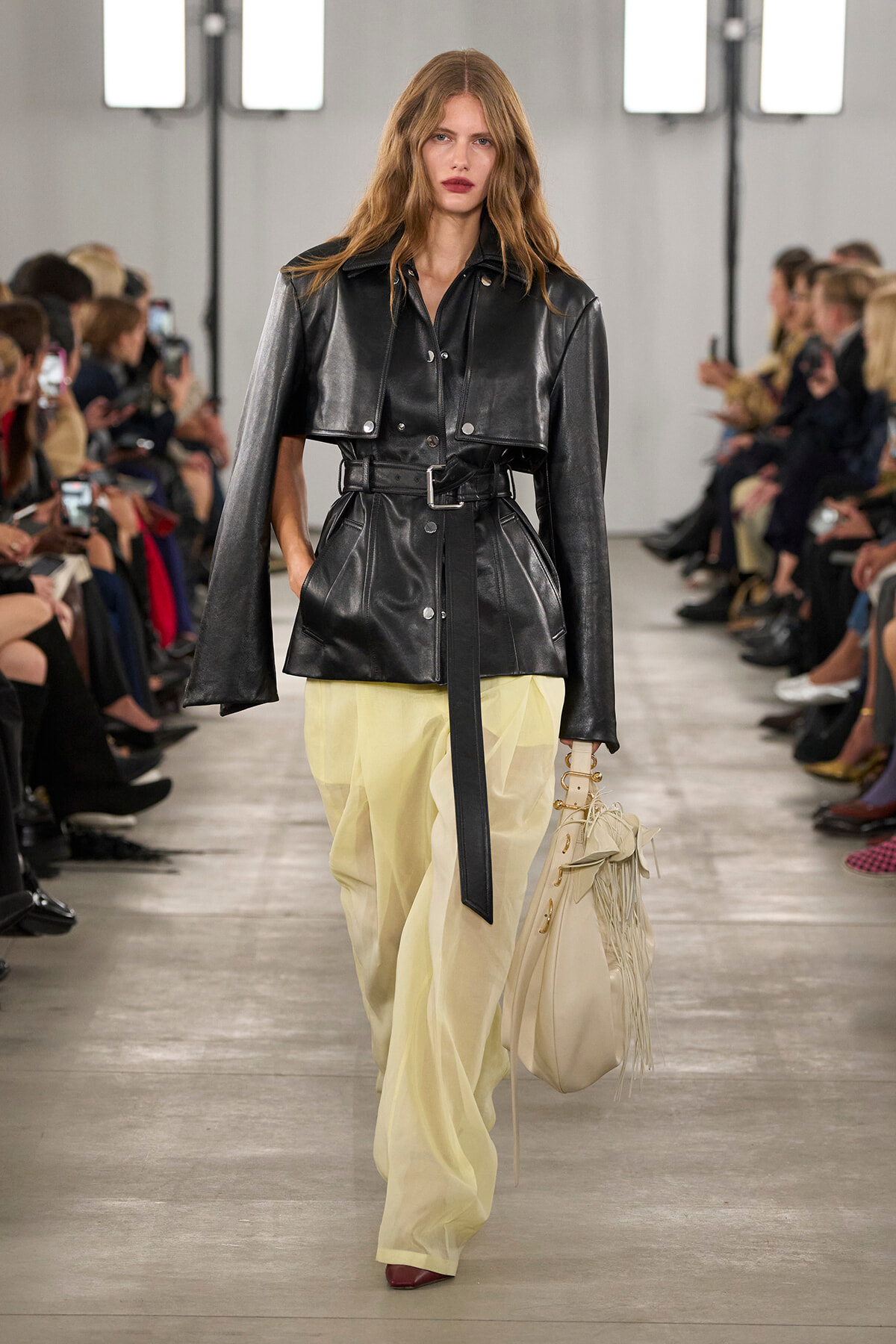 Model walks the runway in a cropped black leather belted jacket with a cape over pale yellow wide-leg pants, carrying a cream fringe bag.