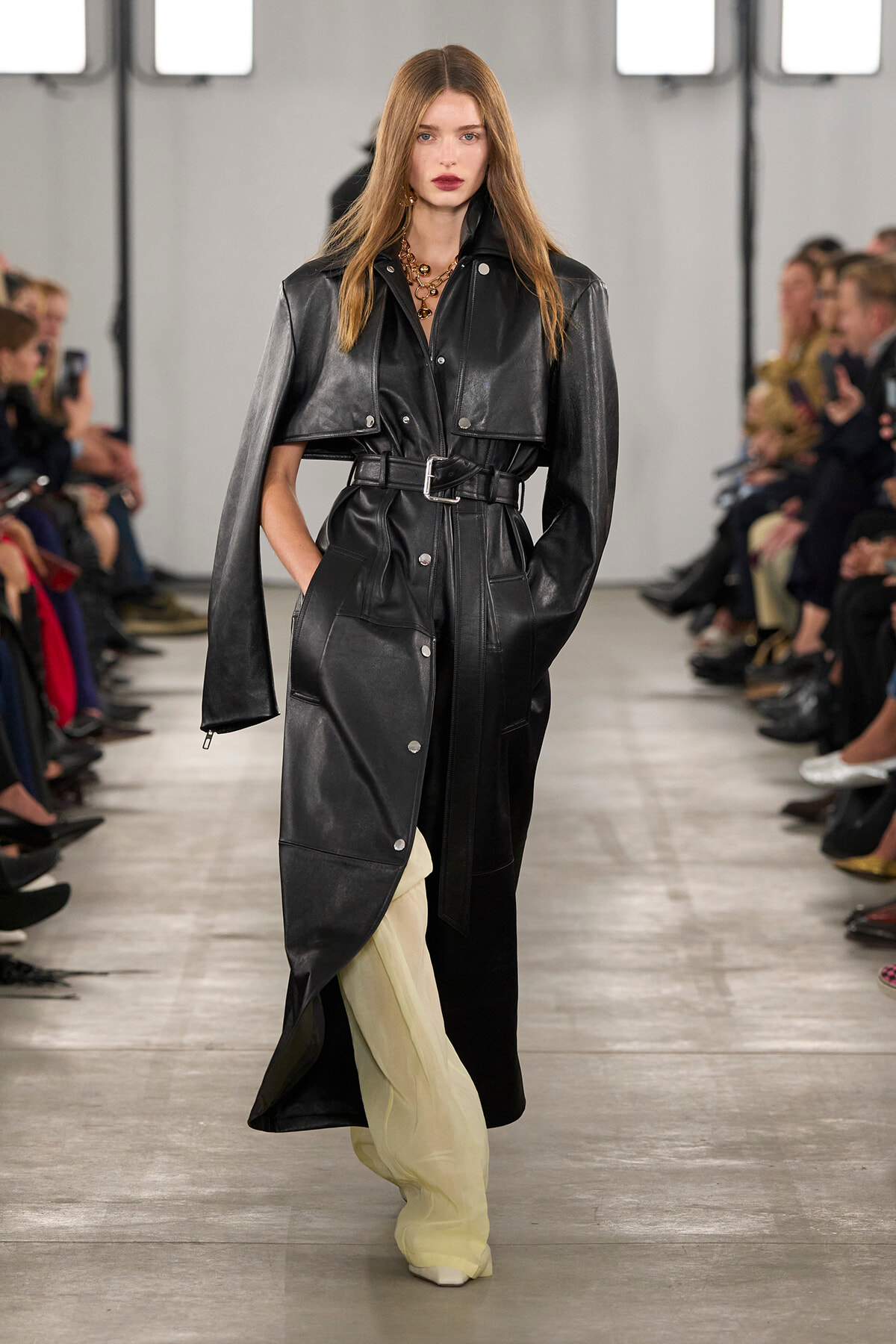 Model walking a runway in a black leather trench coat with a belt, layered over pale yellow pants; audience seated along the sides.