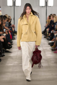 Model walks a runway in a pale yellow structured jacket and wide cream pants, carrying a burgundy drawstring bag with fringe.