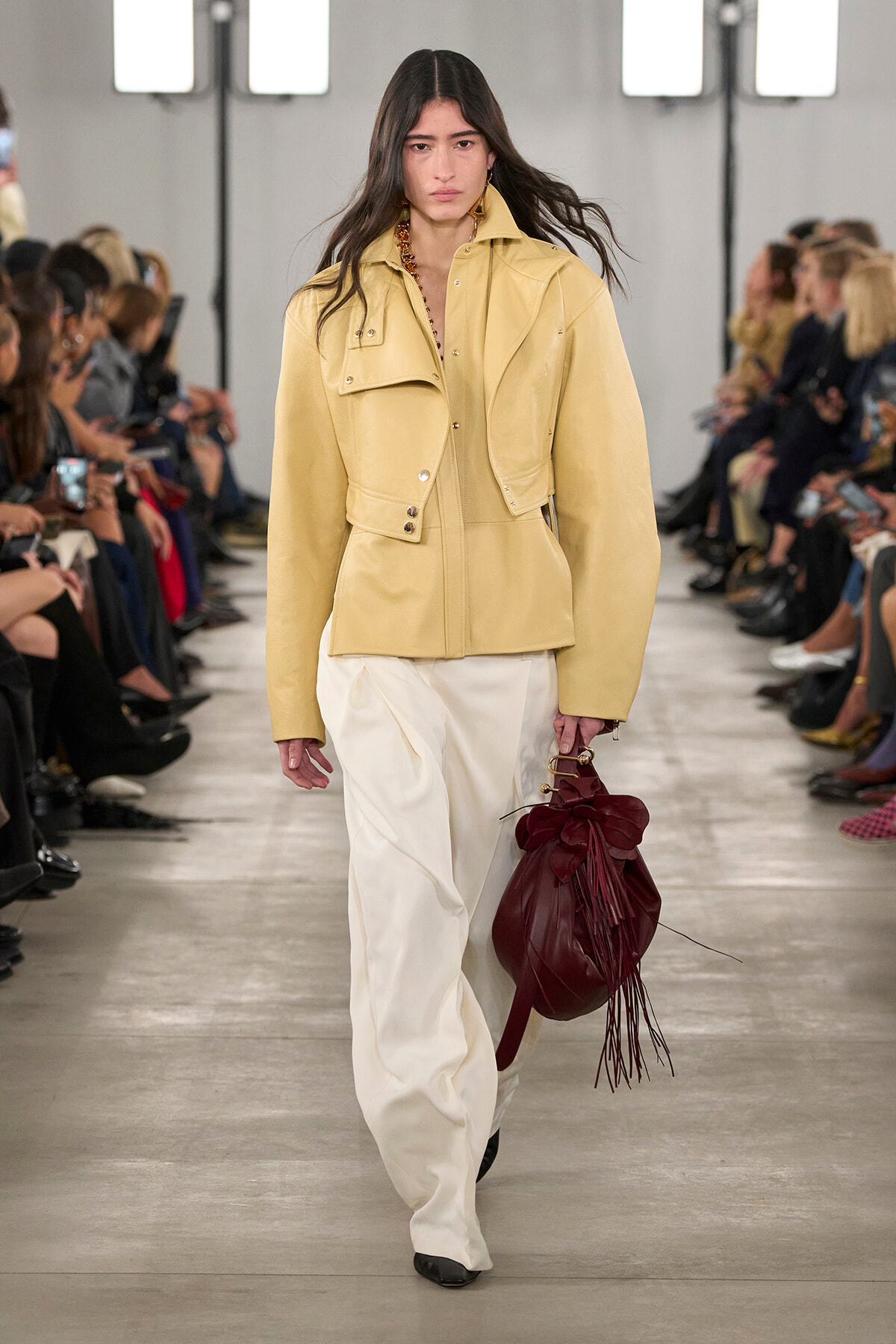 Model walks a runway in a pale yellow structured jacket and wide cream pants, carrying a burgundy drawstring bag with fringe.