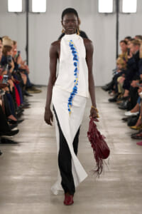 Model walks the runway in a white halter top with a blue feather stripe, paired with white and black wide-leg pants and red shoes, carrying a burgundy fringe bag with gold jewelry nearby.