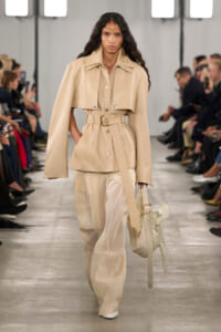 Model walks a runway in an all-neutral beige outfit: oversized cropped jacket with a belt and wide-leg pants, carrying a fringed white handbag.