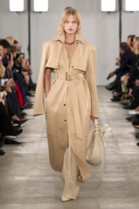 Model walking the runway in a long beige trench with belt, carrying a cream handbag on a fashion show catwalk.