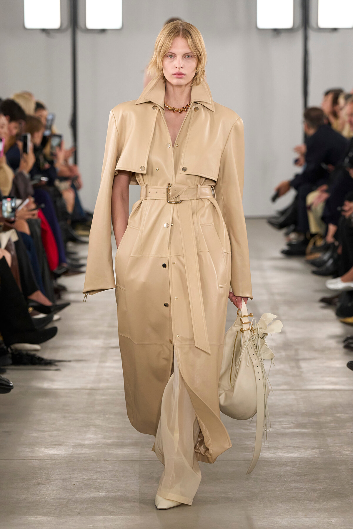 Model walking the runway in a long beige trench with belt, carrying a cream handbag on a fashion show catwalk.
