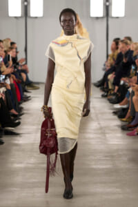 Model walks a runway in a pale yellow cream dress with an abstract print, carrying a burgundy fringe bag as the audience watches.