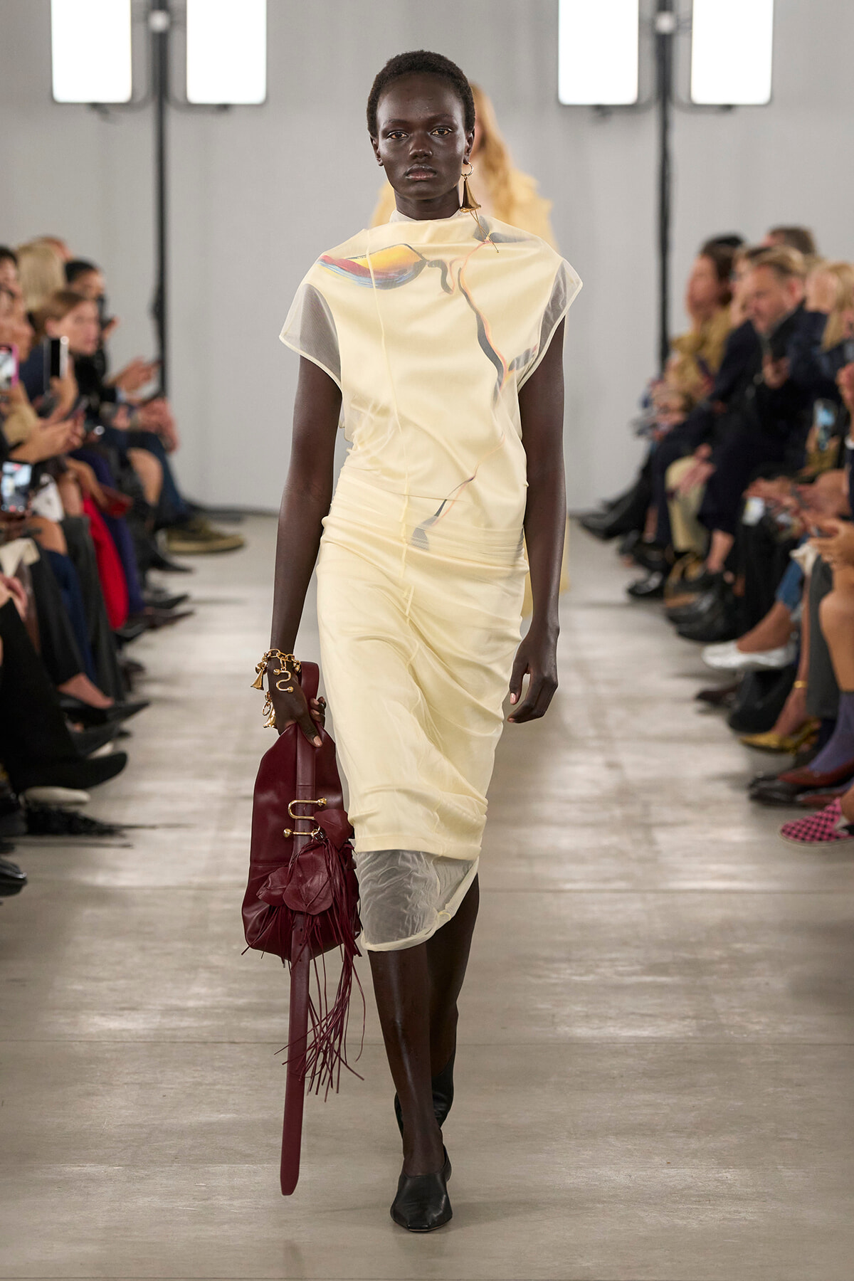 Model walks a runway in a pale yellow cream dress with an abstract print, carrying a burgundy fringe bag as the audience watches.