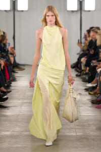 Model walking the runway in a pale yellow halter-neck gown with a slit-hem sheer underlayer, carrying a beige tassel handbag on the right.