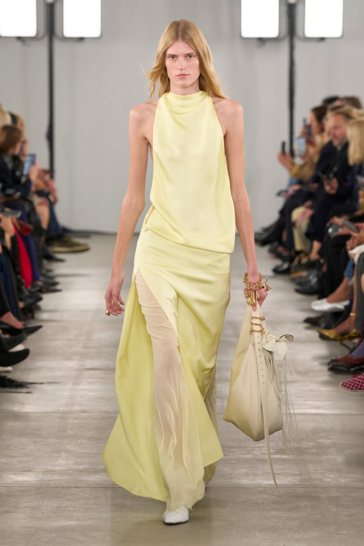 Model walking the runway in a pale yellow halter-neck gown with a slit-hem sheer underlayer, carrying a beige tassel handbag on the right.