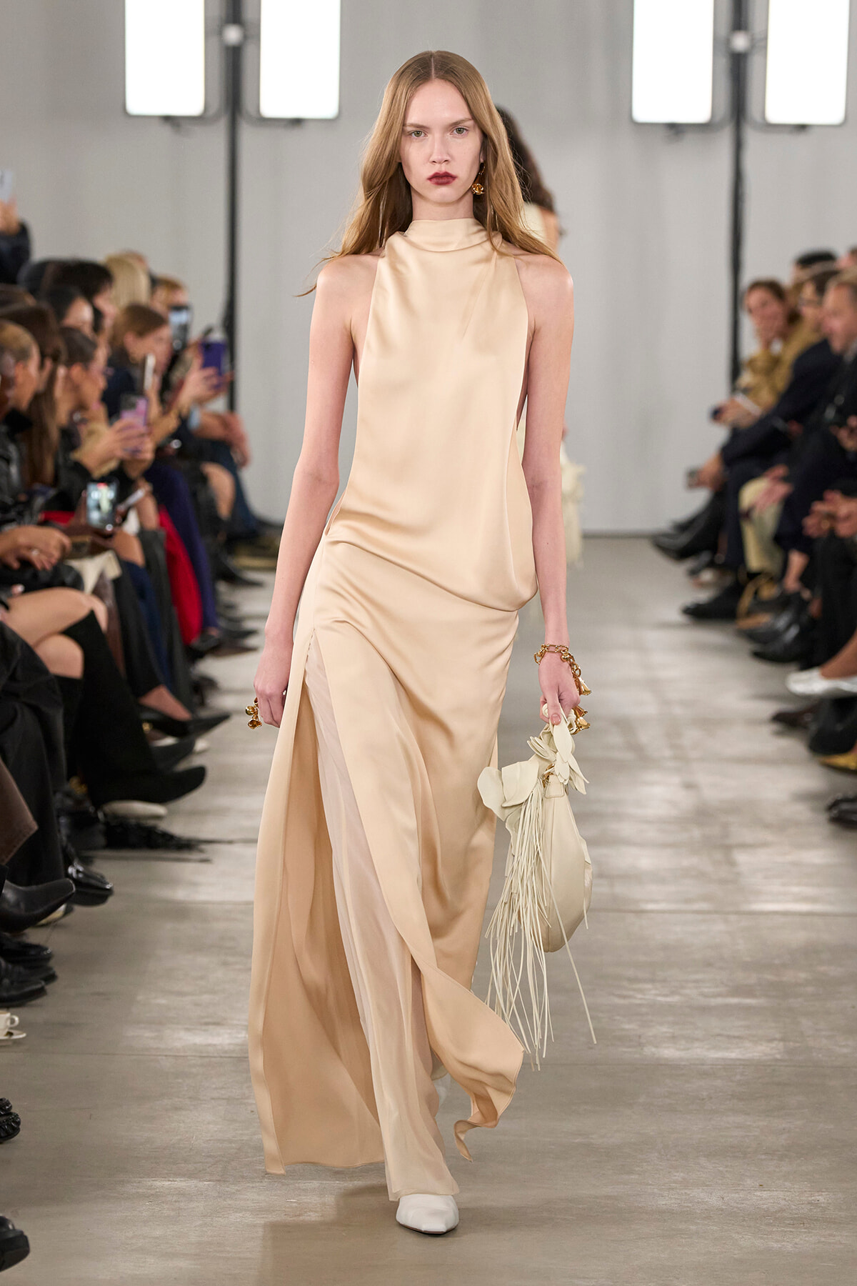 Model walks a fashion show in a beige satin halter-neck gown with a flowing skirt, carrying a fringed bag; audience watches.