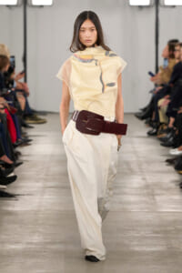 Runway model wearing a cream abstract-print top, white wide-leg pants, and a thick burgundy belt, walking the catwalk.