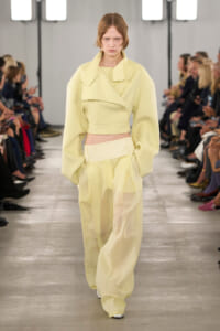 Model walking a fashion runway in a pale yellow oversized jacket paired with wide-leg pants, audience on both sides watching.