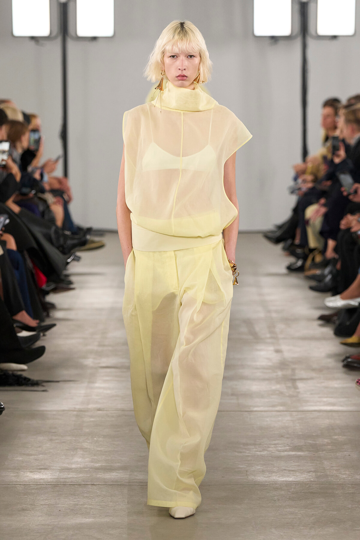 Model walking a fashion runway in a pale yellow sheer top and wide-leg pants, with an audience watching from both sides.