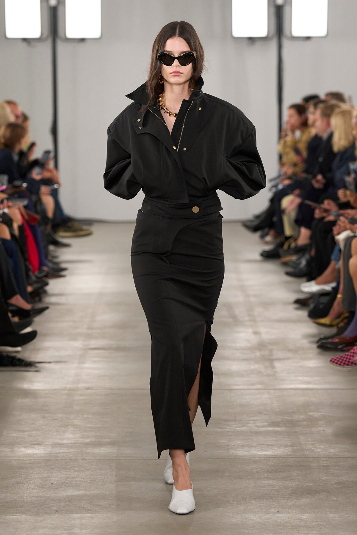 Model walking a fashion runway in an all-black ensemble: oversized jacket, high-waisted skirt with a front slit, white slip-on shoes, cat-eye sunglasses, and a chunky gold necklace.