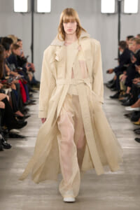 Model walking the runway in a long beige trench coat with belt, sheer inner layers, and white shoes.