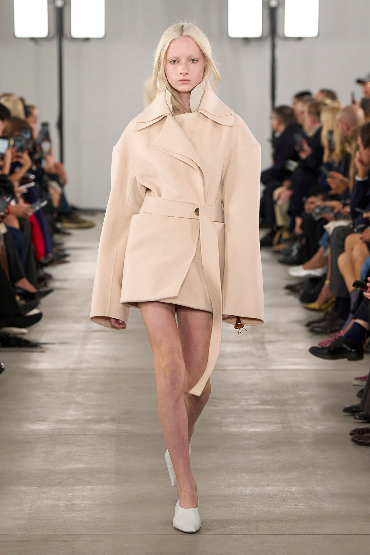 Model walking a runway in a pale beige oversized wrap coat with a belt and wide collar, white heels, audience in the background, neutral expression.