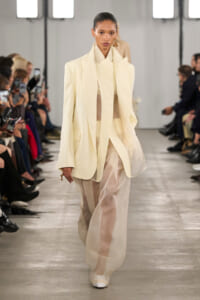 Model walks a fashion runway in a cream oversized blazer and sheer pale trousers, audience photographing the show in the background.
