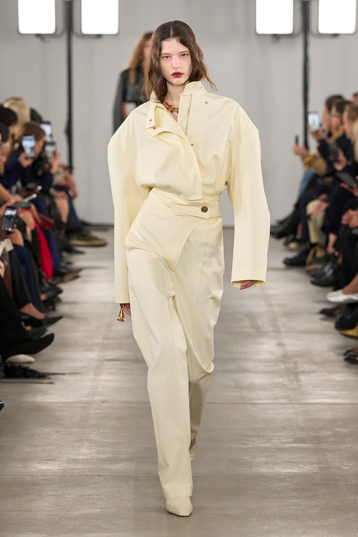 Model walks a cream tailored suit down a runway, oversized jacket and high-waisted pants, audience filming on both sides.
