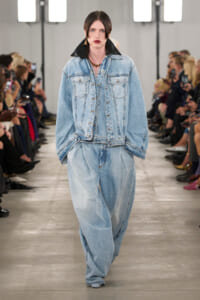 Model walks the runway in an oversized light-wash denim jacket with a black collar and baggy matching jeans; audience lines the catwalk.