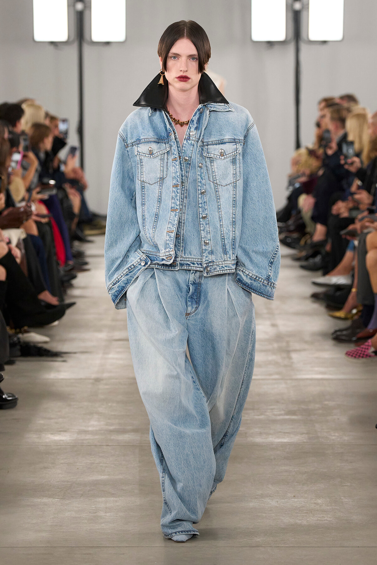 Model walks the runway in an oversized light-wash denim jacket with a black collar and baggy matching jeans; audience lines the catwalk.