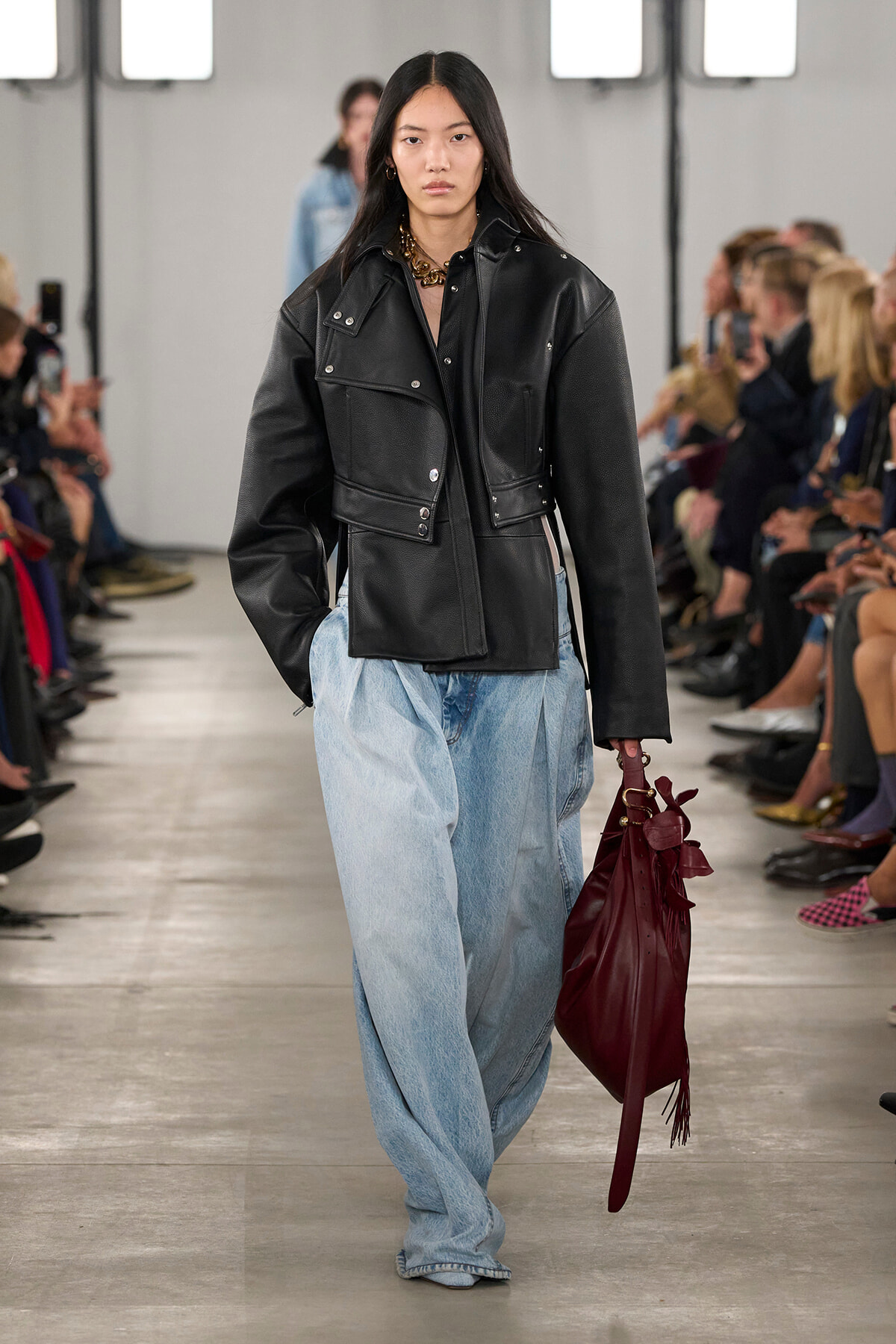 Model walks a fashion runway in an oversized black leather jacket and wide light-blue jeans, carrying a burgundy handbag.