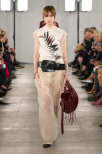 Model on a runway wearing sheer beige wide-leg pants, a white top with a black leaf print, a thick black belt, and a burgundy fringed bag.
