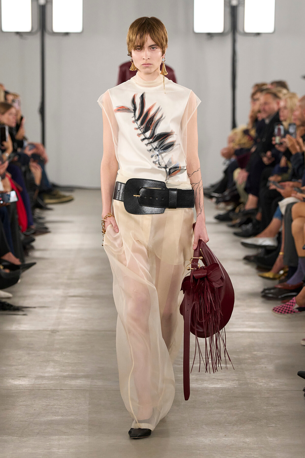Model on a runway wearing sheer beige wide-leg pants, a white top with a black leaf print, a thick black belt, and a burgundy fringed bag.