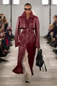 Model walks the runway in a burgundy leather trench coat with a matching belt, oversized collar, and a black fringed bag paired with dark sunglasses.