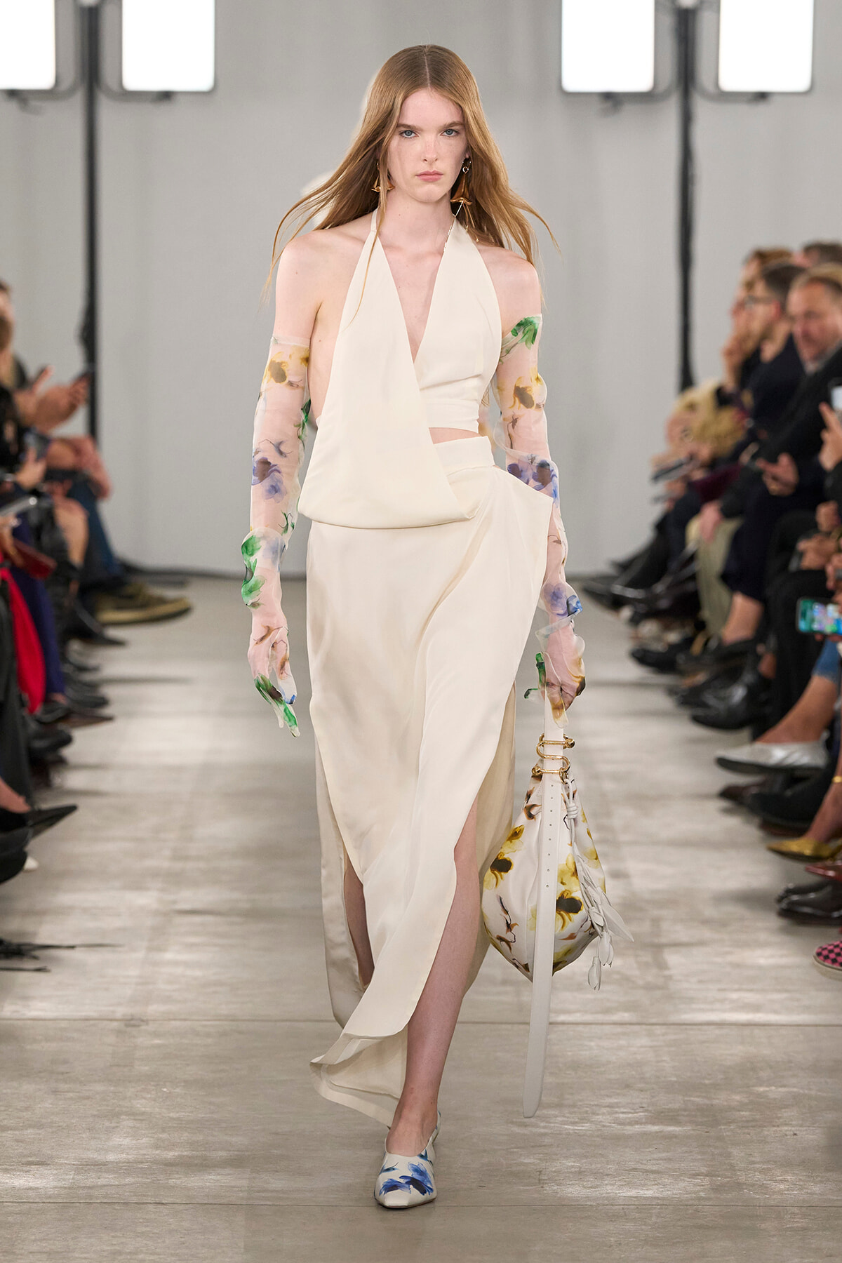 Model walking a runway in a cream halter gown with a draped top and high slit, paired with floral sheer sleeves and a matching floral bag.