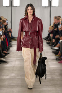 Female model on a runway wearing a burgundy leather belted jacket with a wide collar and matching belt, paired with cream wide-leg pants and a black fringed bag