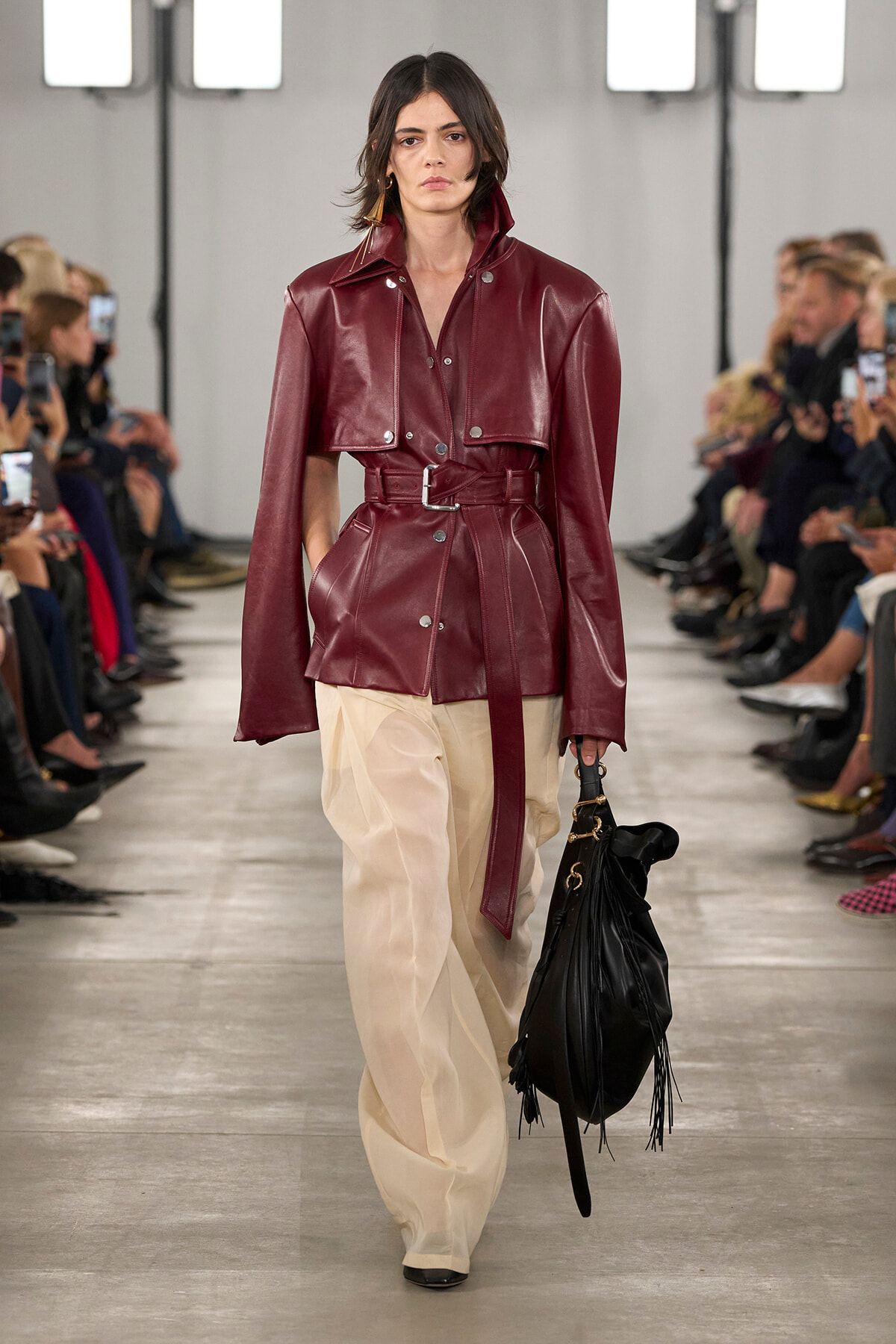 Female model on a runway wearing a burgundy leather belted jacket with a wide collar and matching belt, paired with cream wide-leg pants and a black fringed bag
