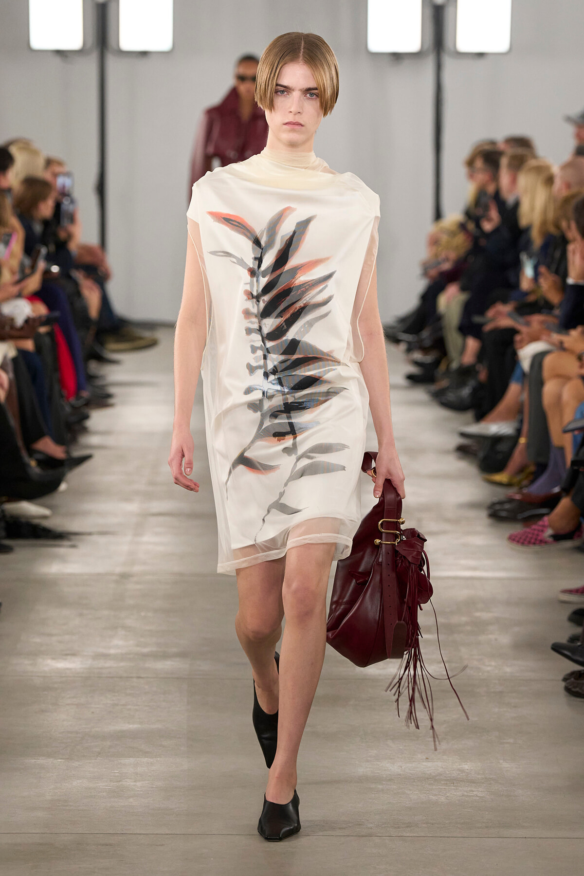 Model walking a runway in a white knee-length dress with a bold leafy print, carrying a burgundy fringe bag and wearing black mules.