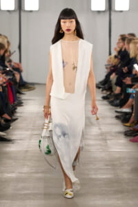 Female model walks a fashion runway in a white sleeveless vest over a sheer floral top and a white gradient-print skirt, carrying a white handbag with green leaf design.