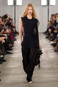 Model walks a black, sleeveless tunic with sheer panels and wide black trousers down a runway, carrying a fringed bag.