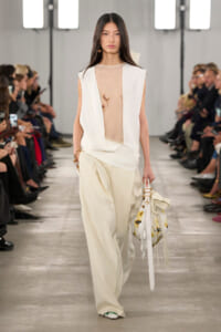 Model walks the runway in an ivory sleeveless vest over a sheer top and wide-leg pants, carrying a white floral handbag with fringe.