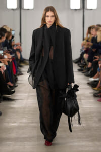 Model walks a runway in an oversized black blazer over sheer black pants, carrying a black fringed bag and wearing red shoes.