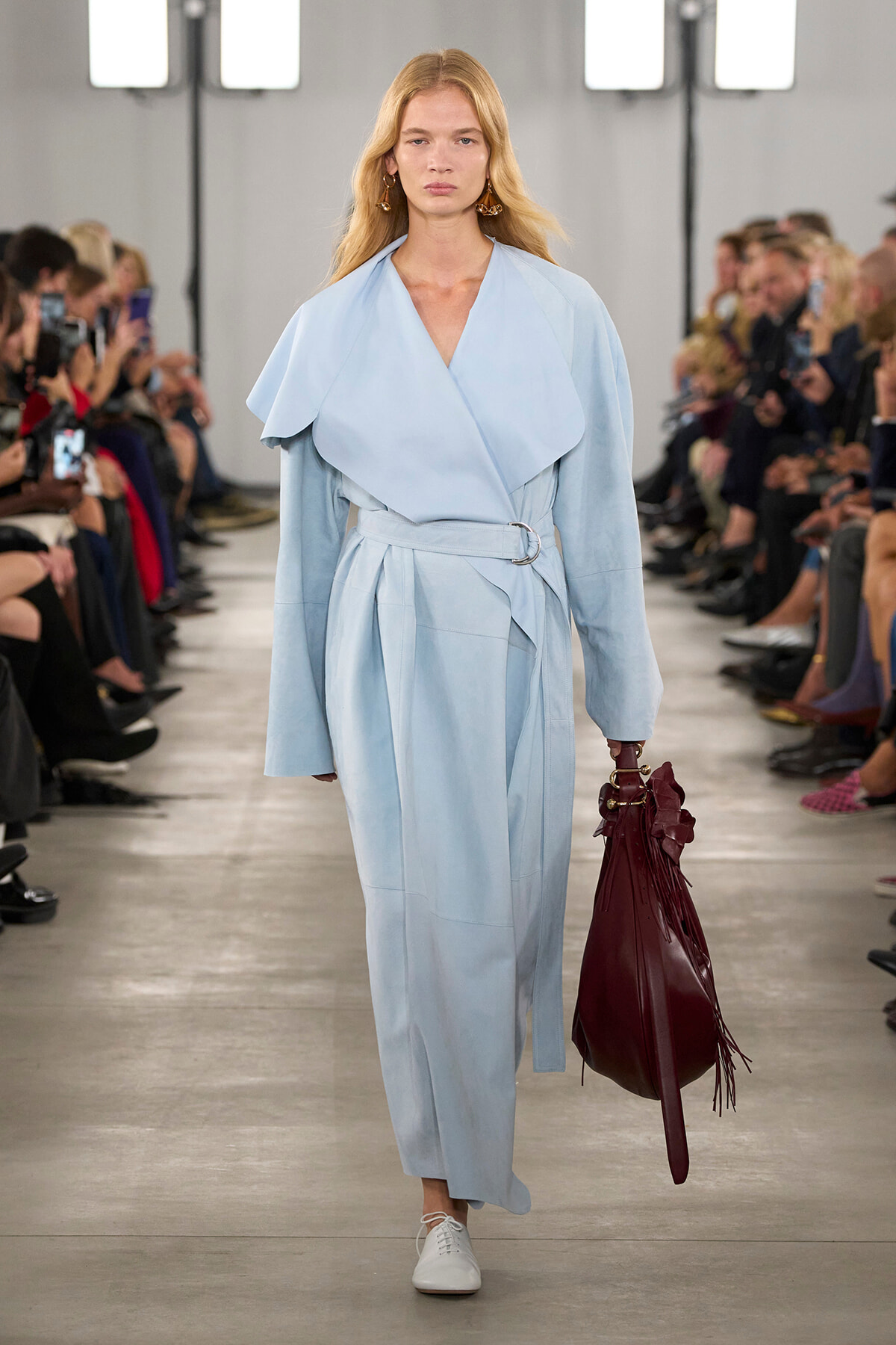 Model walks a runway in a pale blue wrap coat with a belt, white sneakers, and a large burgundy fringe handbag; audience watches from both sides.