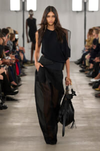Model walking a runway in an all-black outfit with a draped one-shoulder top and sheer wide-leg pants, carrying a black fringe bag.
