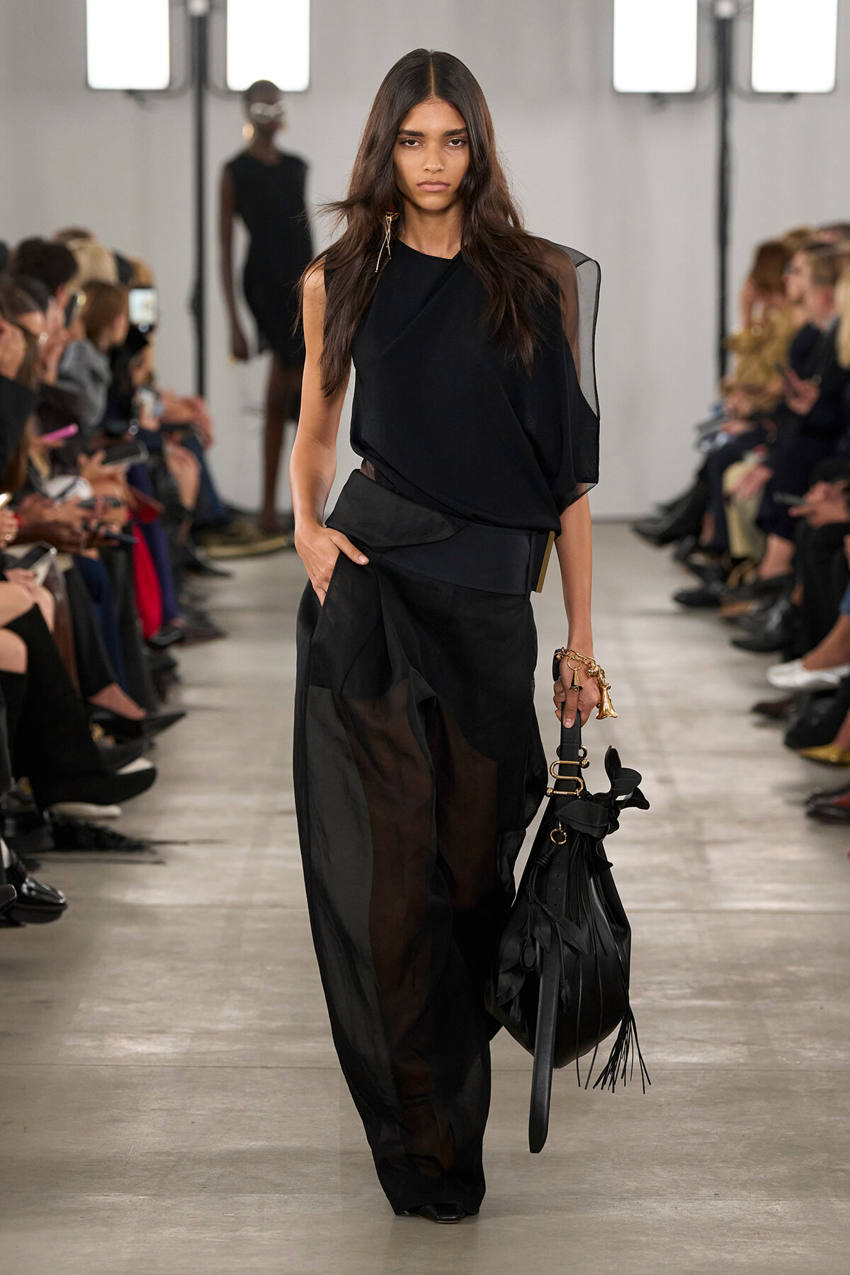 Model walking a runway in an all-black outfit with a draped one-shoulder top and sheer wide-leg pants, carrying a black fringe bag.