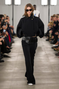 Model walks a minimalist black ensemble on a runway, wearing a loose black top, wide belt, flared pants, and dark sunglasses.