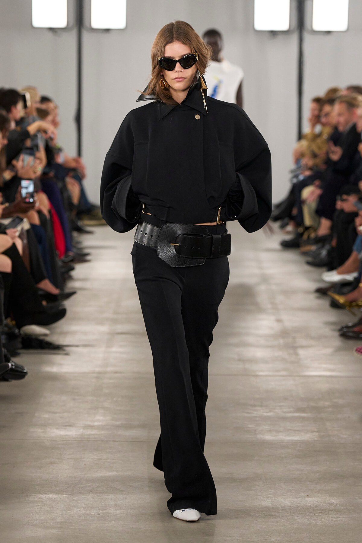 Model walks a minimalist black ensemble on a runway, wearing a loose black top, wide belt, flared pants, and dark sunglasses.
