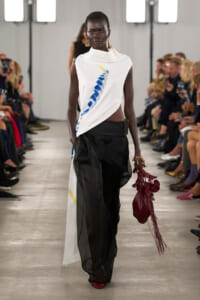 Model walks a fashion runway in a white draped sleeveless top with a blue feather design, sheer black pants, and a burgundy fringe bag in hand amid seated audience.