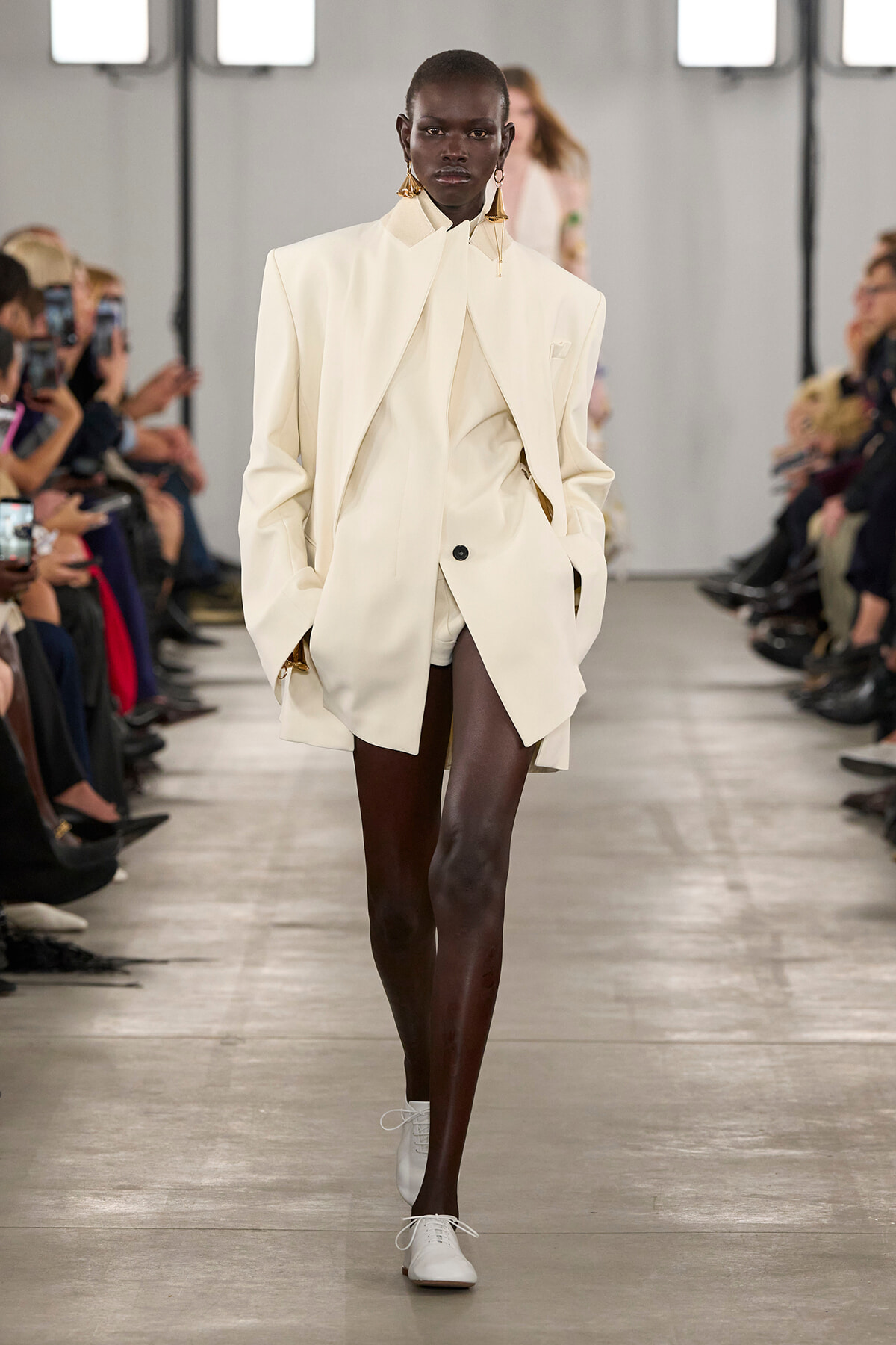 Black female model on a runway wearing an oversized ivory blazer with matching shorts and white lace-up shoes.