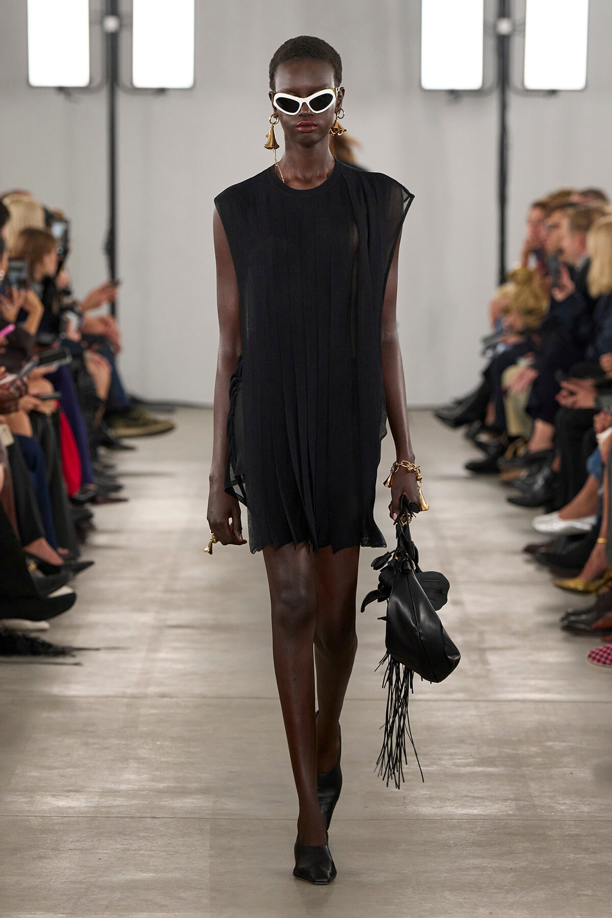 Model on a runway wearing a black sleeveless pleated dress, white cat-eye sunglasses, and gold earrings, carrying a black fringed bag.