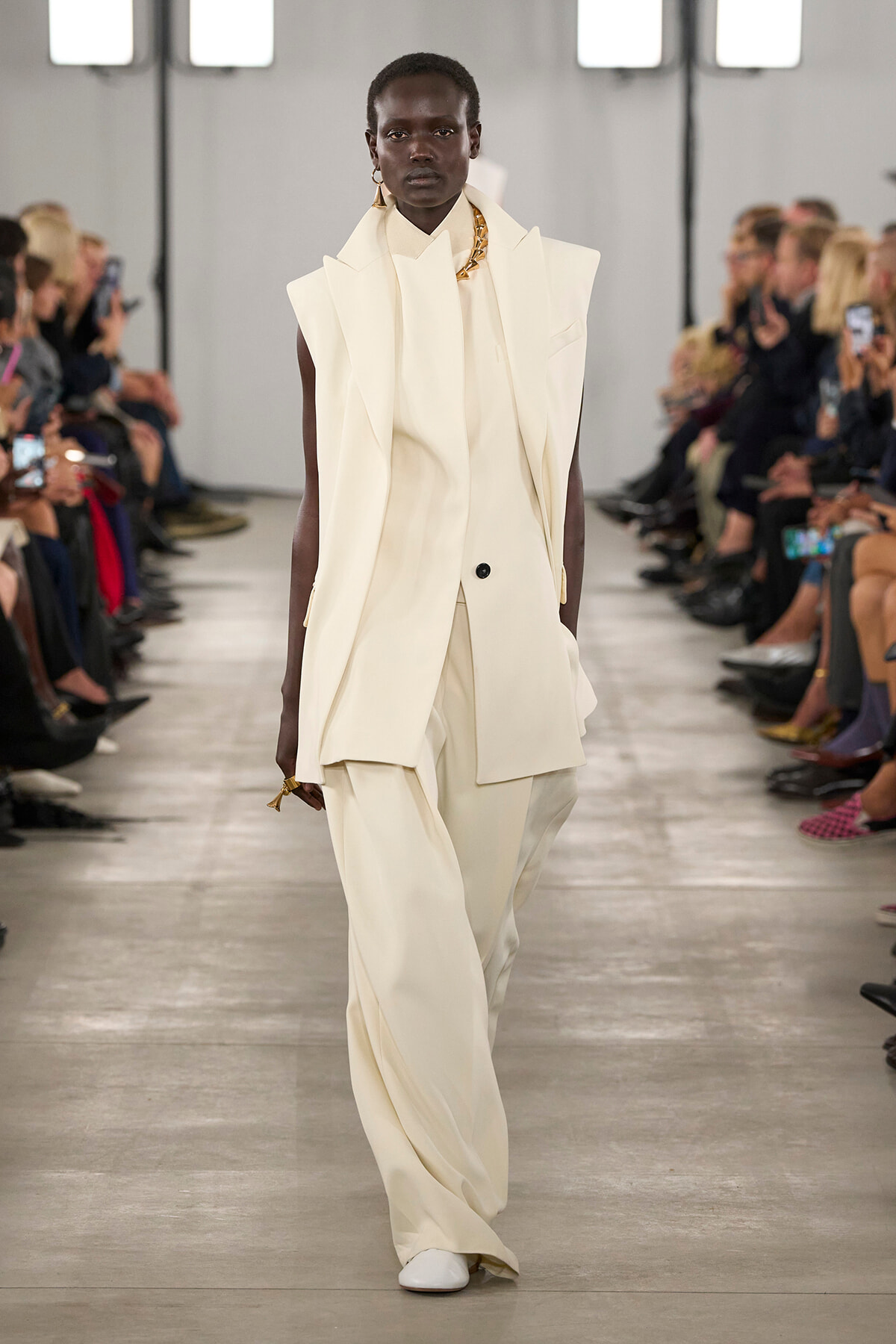 Model walks the runway in an oversized cream suit with layered vest and wide-leg pants, gold jewelry visible, audience blurred in background.