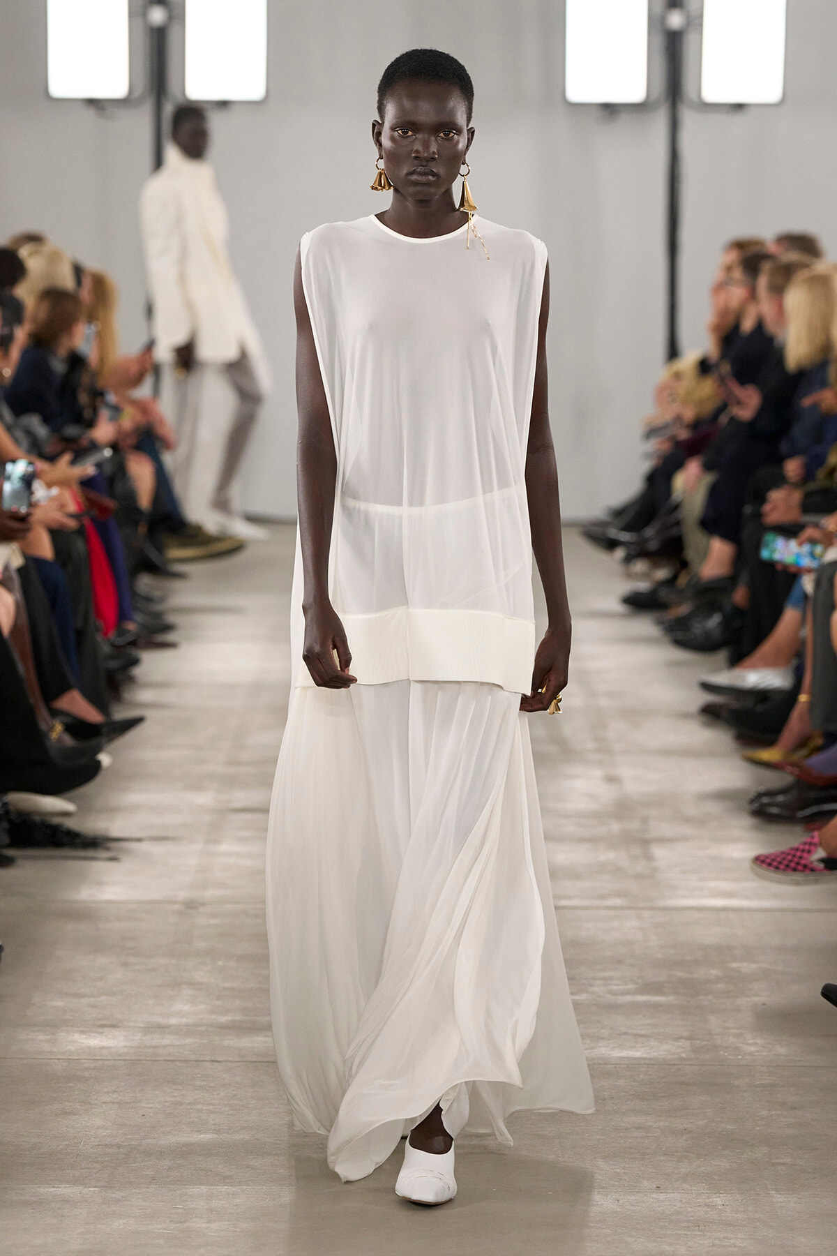 Model walks a black woman down the runway in a flowing sheer white sleeveless dress with a two-tier hem and white shoes, wearing large gold earrings.