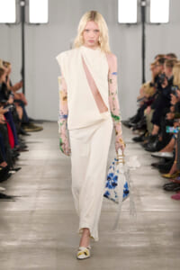 Model walks a runway in an ivory asymmetrical dress with a diagonal sheer panel, paired with floral long gloves; she carries a blue-and-white floral handbag with a fringe and wears matching floral shoes.