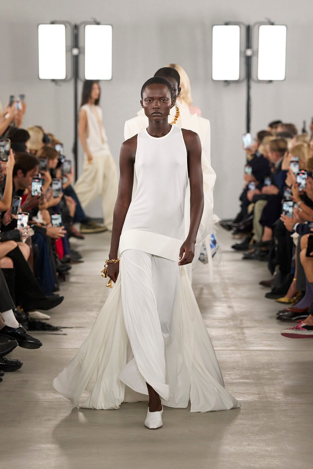 Black model walks a white sleeveless gown down the runway, with a long flowing train, white shoes, and gold bracelets as photographers watch from both sides under bright studio lights.