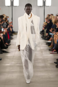 Model walks a runway in an oversized white suit with a sheer inner layer and gold chain necklace, audience on both sides watching.